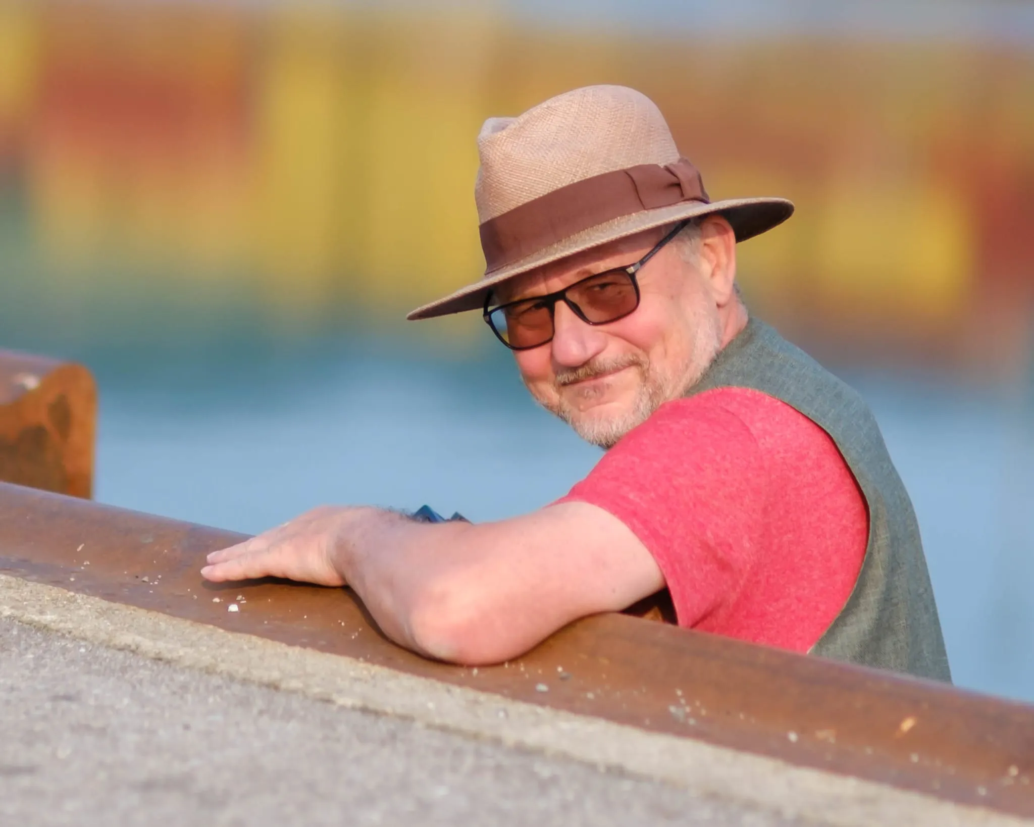 Mann mit hellem Hut und Sonnenbrille lehnt an einem rostigen Geländer, dahinter Wasser