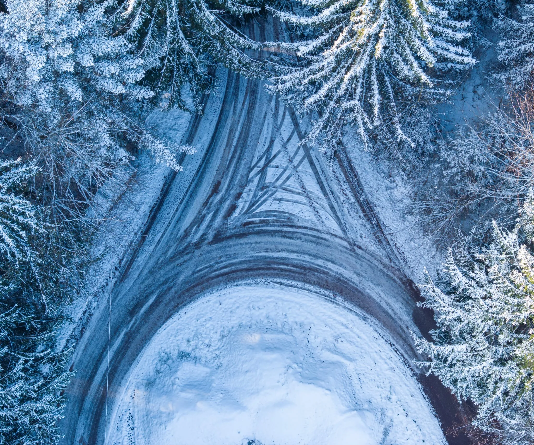 Senkrechte Luftaufnahme einer schneebedeckten Straßenkurve, gesäumt von verschneiten Nadelbäumen
