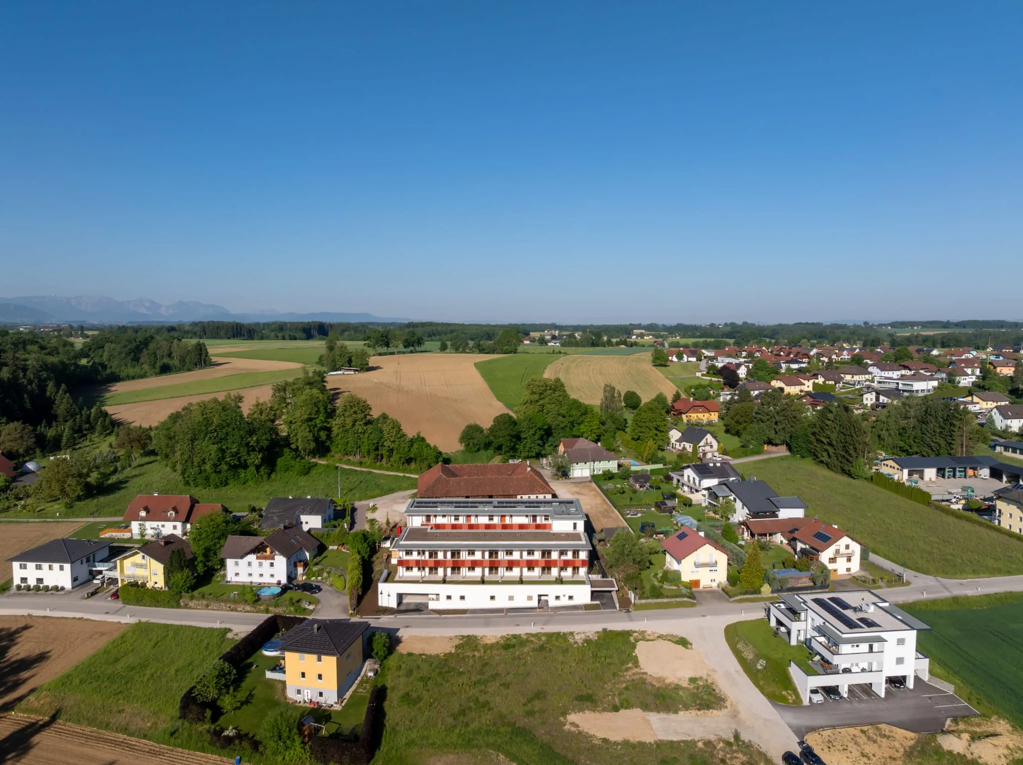 Luftaufnahme einer Ortschaft mit Feldern und Wiesen, im Zentrum eine moderne Wohnanlage, am Horizont Berge