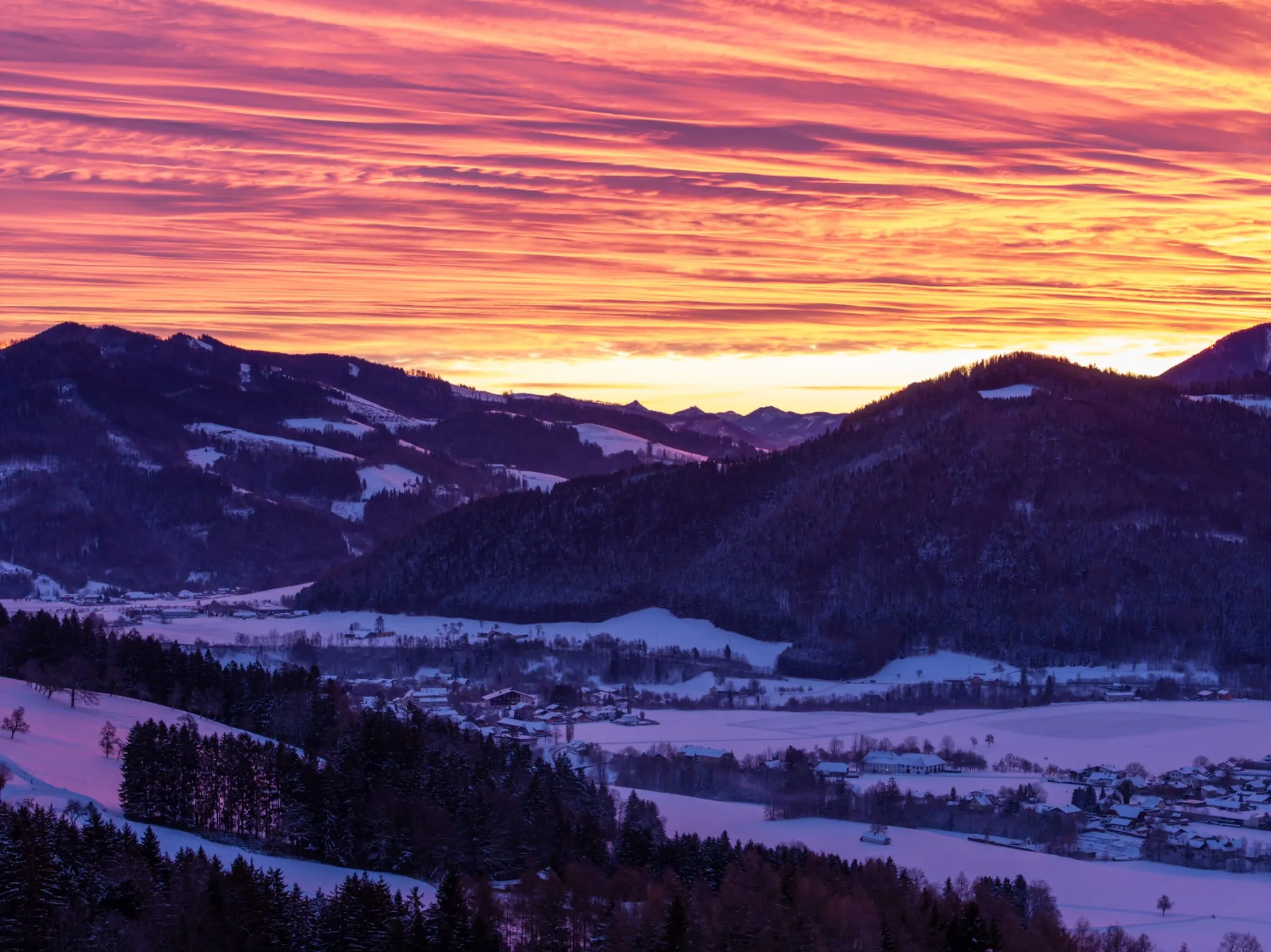 Verschneites Tal unter einem rot-violett gefärbten Himmel über bewaldeten Hügeln