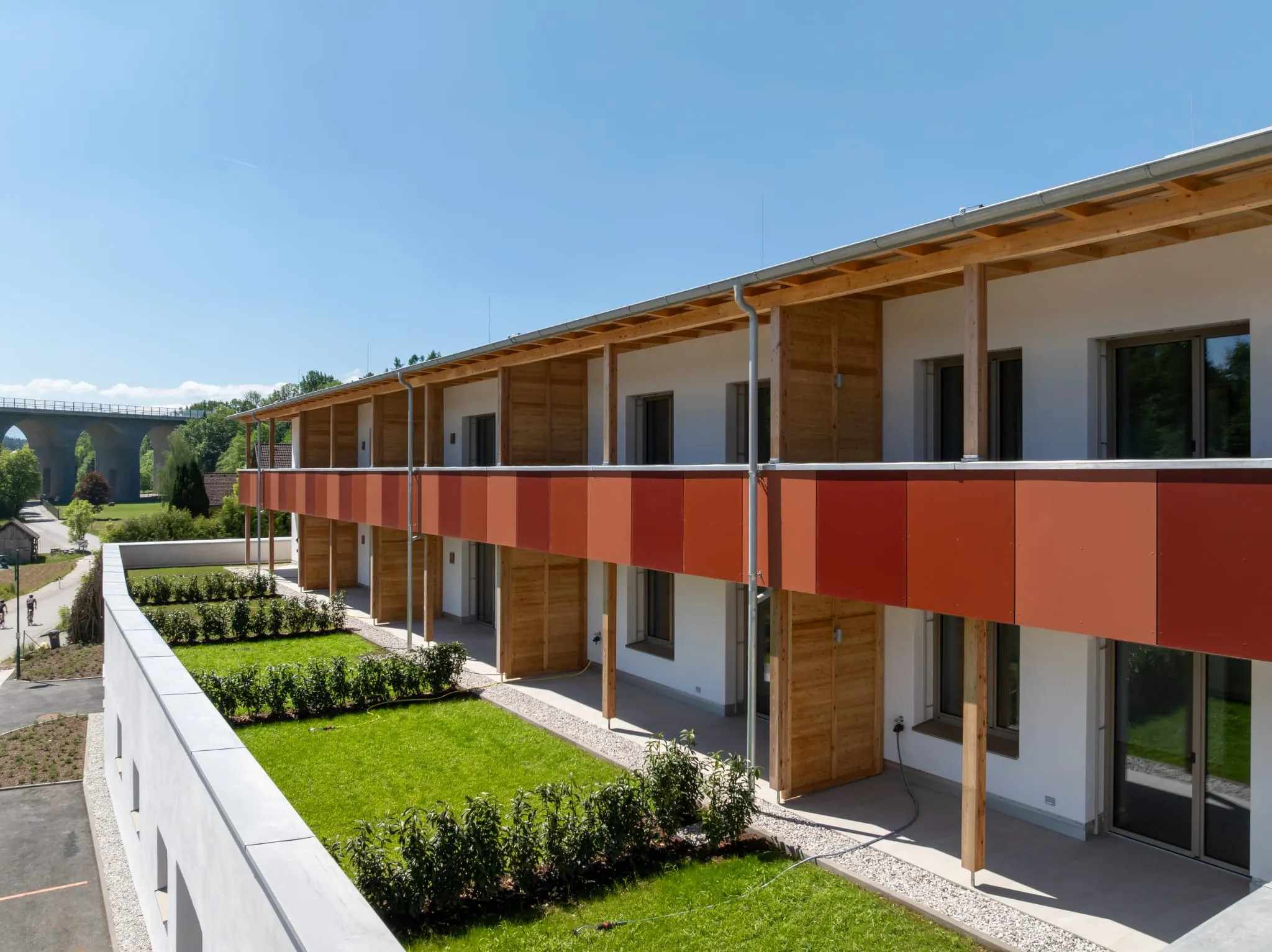 Moderne zweigeschossige Wohnanlage mit orangefarbenen Balkonpaneelen und Holzfassade, davor frisch angelegte Beete