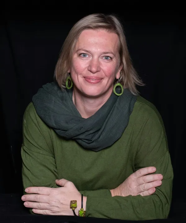 Die Fotografin Manuela Rathberger im grünen Pullover mit dunkelgrünem Schal, die Arme verschränkt, vor dunklem Hintergrund